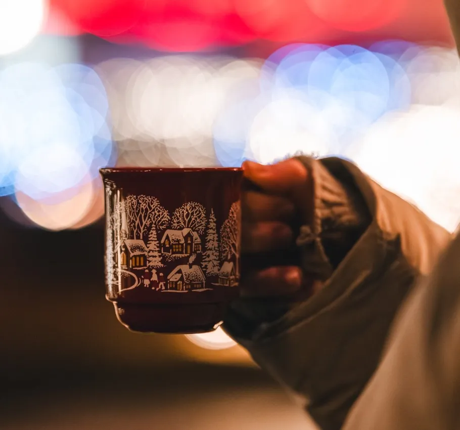 Deutsch   Weihnachtliche Tasse mit Heißgetränk in festlicher Atmosphäre (Titel)  Person hält eine weihnachtlich verzierte Tasse mit Heißgetränk vor stimmungsvollen Lichtern und festlicher Weihnachtsbeleuchtung.