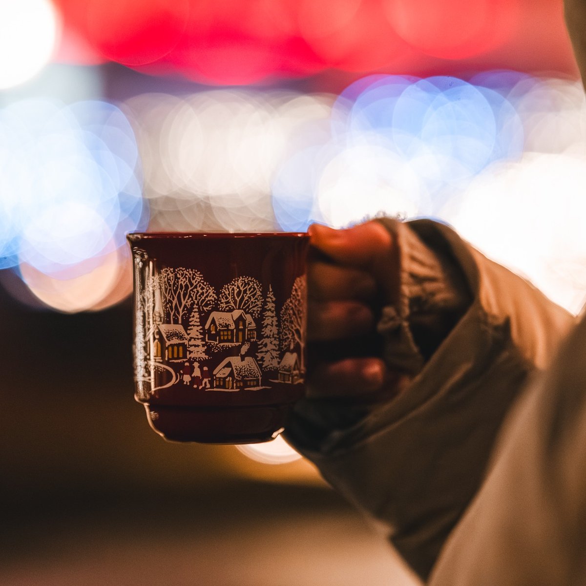 Person holding a festive Christmas mug filled with a hot drink against a background of warm holiday lights and seasonal decorations.